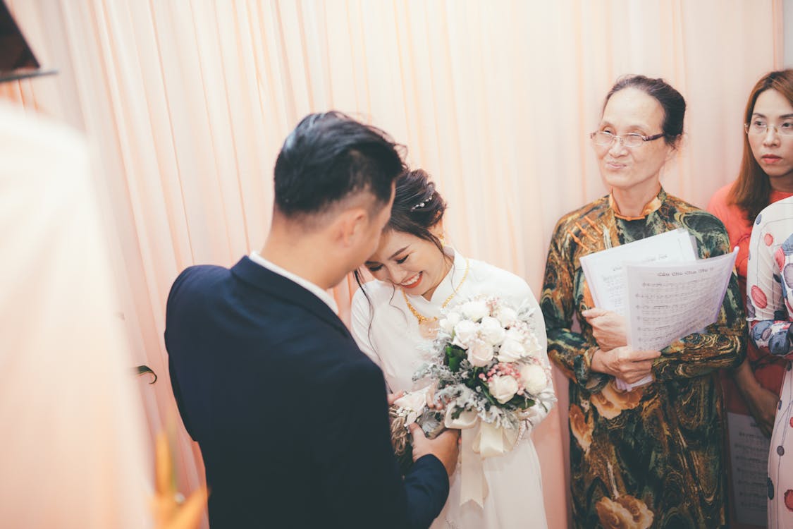 Bride smiling at groom during a wedding ceremony, with an elderly woman holding documents in the background.