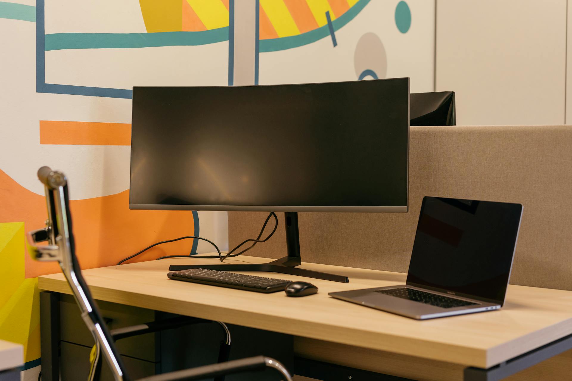 A modern office setup featuring a sleek computer monitor and a laptop on a wooden desk with a colorful abstract background.