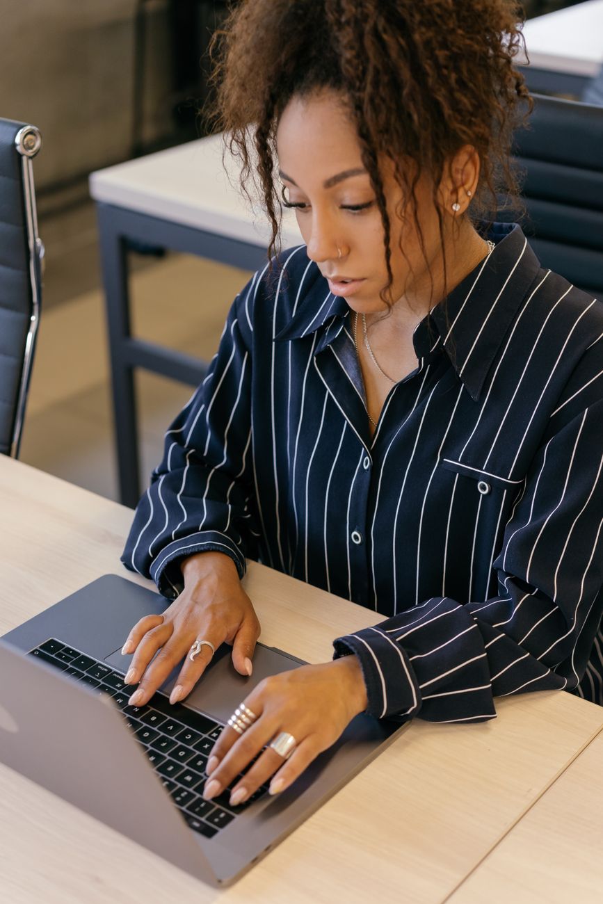 An African American woman with curly hair working on a laptop in an office setting, wearing striped long sleeves.