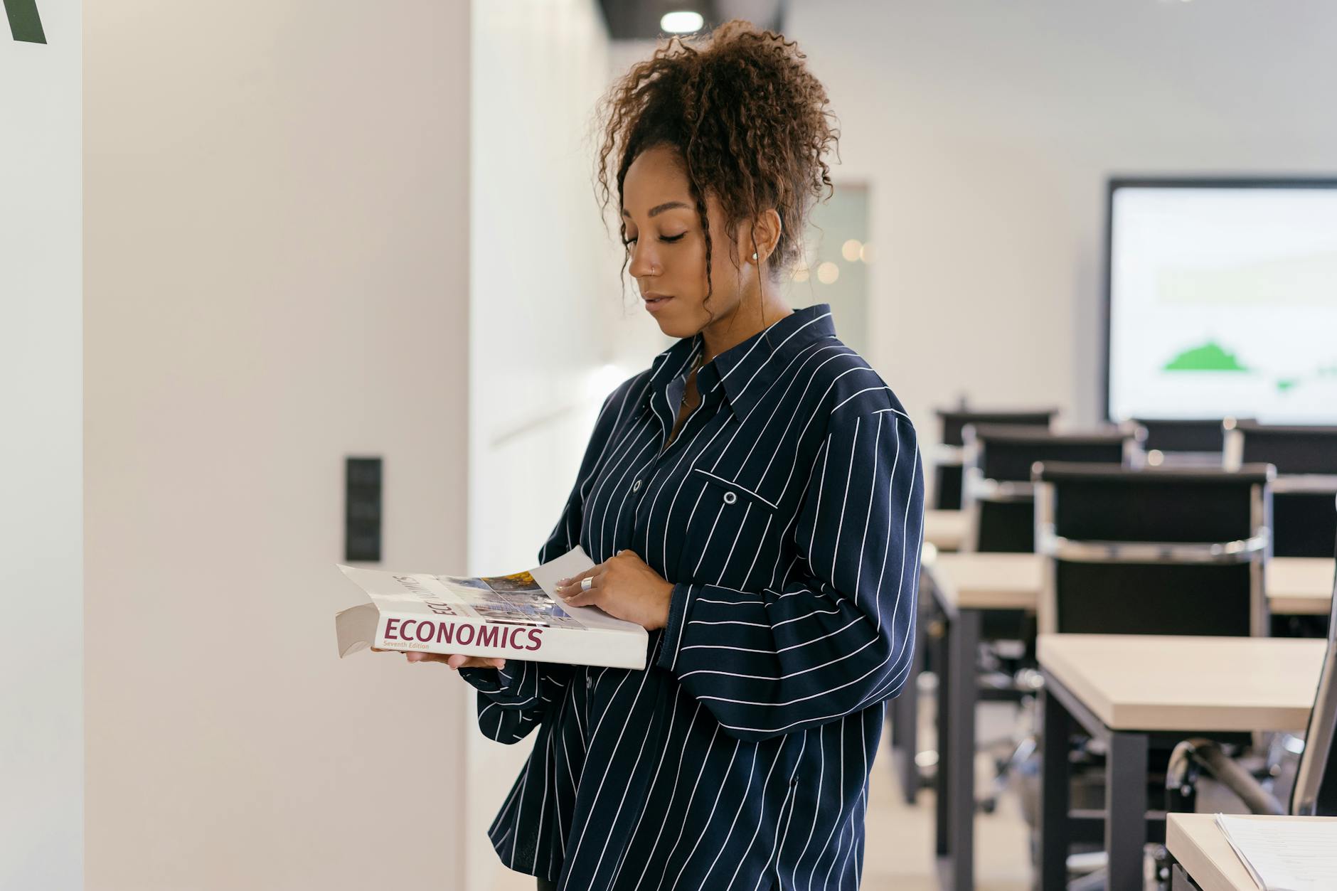 Young woman with curly hair reading economics book in a modern classroom.