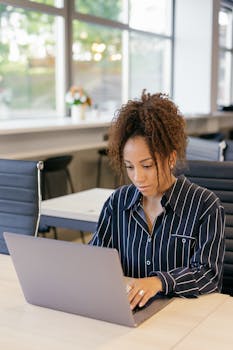 Young African American woman concentrating on her work at a laptop in a bright, modern office space.