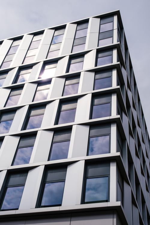 Free A striking modern office building facade with reflective glass windows and geometric patterns under a white sky. Stock Photo