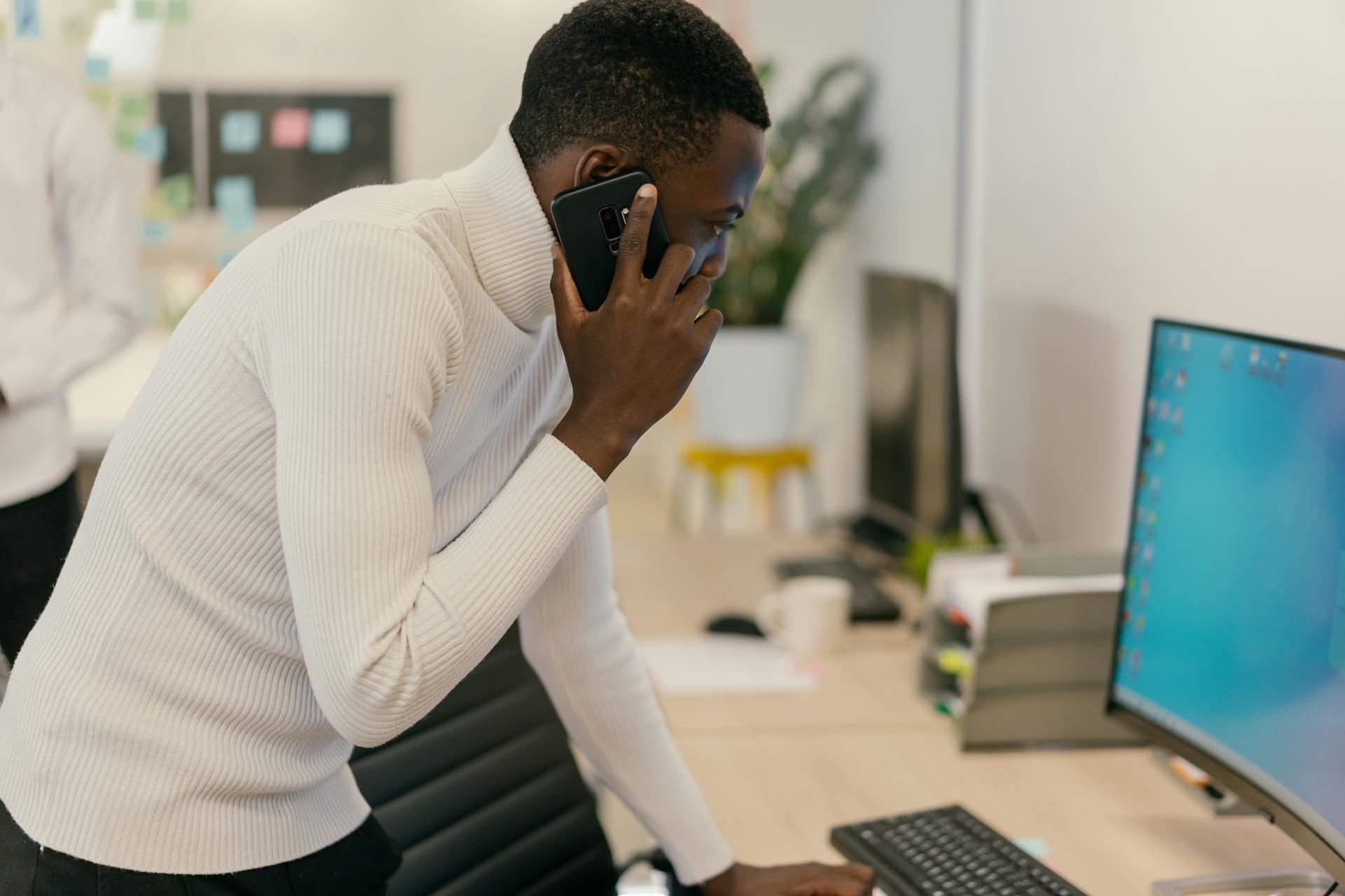 Businessman in white turtleneck talks on smartphone while working at a desk.