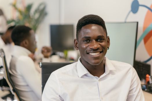 A cheerful young man in a white shirt smiling at his desk in a modern office environment.