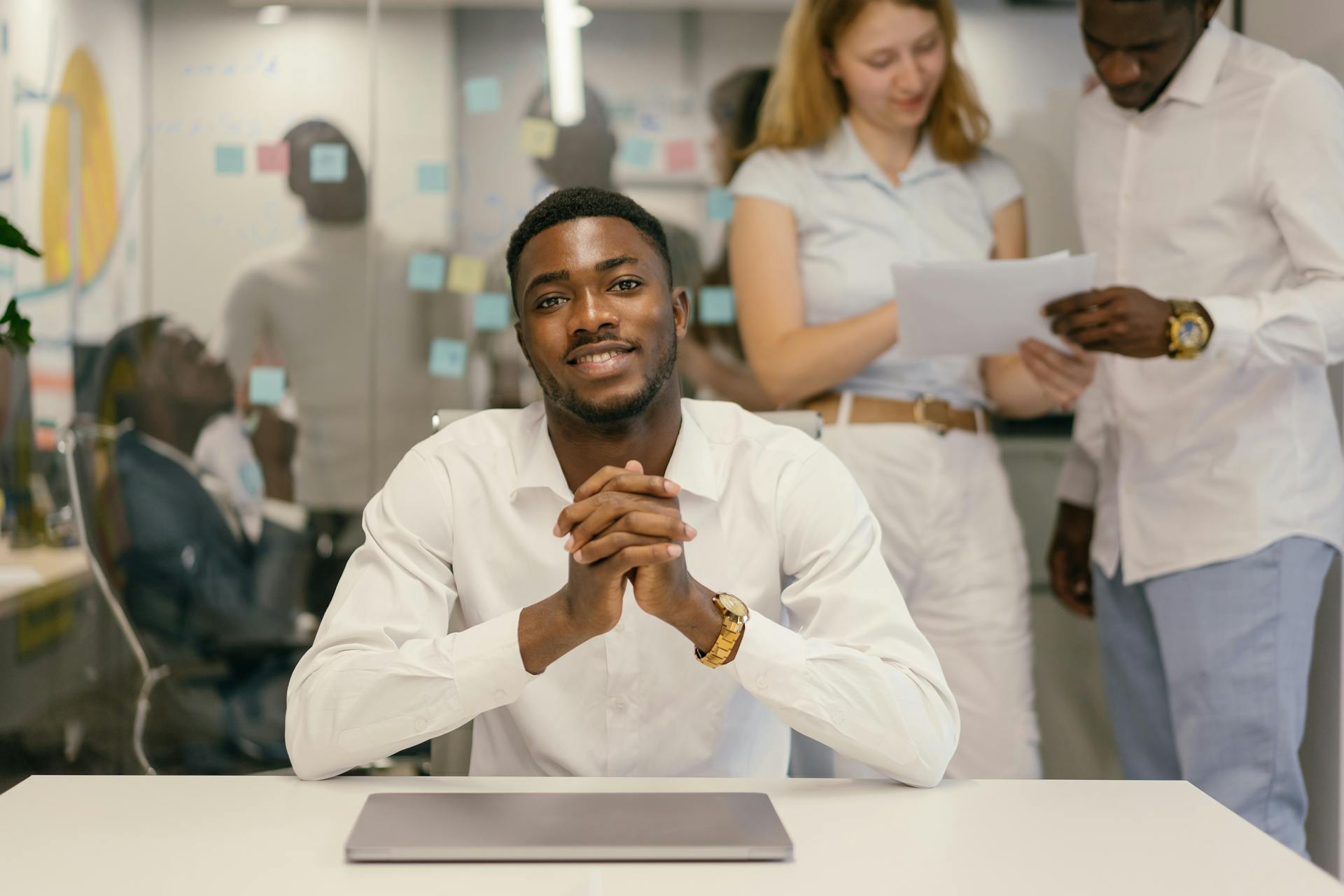 Confident businessman seated at a desk in a collaborative office environment with colleagues in the background.