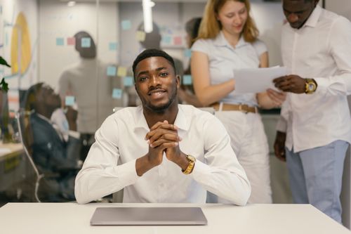 Free Confident businessman seated at a desk in a collaborative office environment with colleagues in the background. Stock Photo