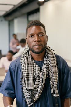 A confident man wearing a blue shirt and checkered scarf poses in an office environment, suggesting professionalism.