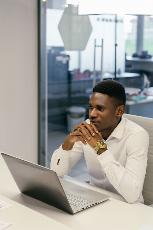 Free African man in white shirt focused on laptop in a contemporary office setting, hands folded. Stock Photo