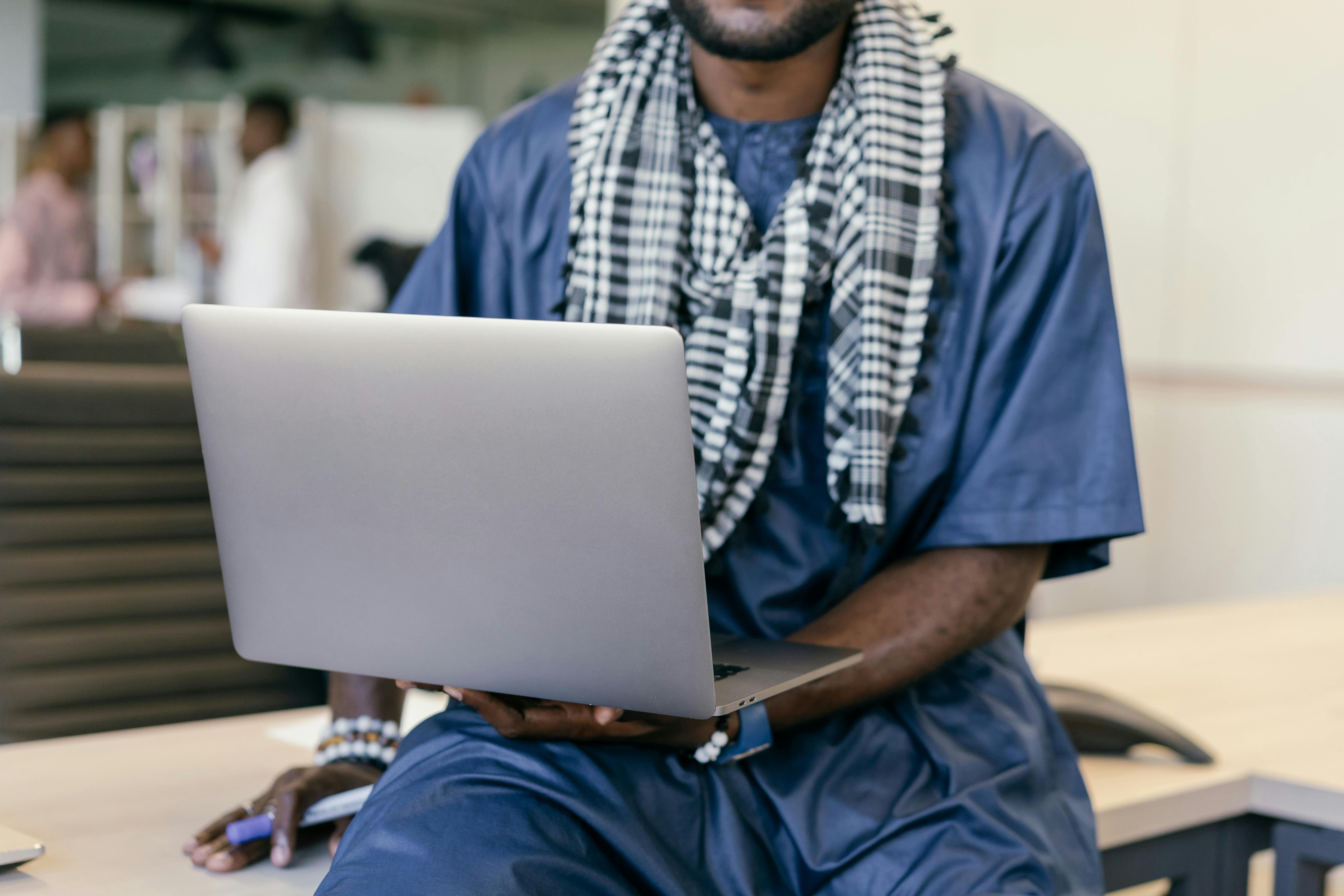 African man in traditional clothing using a laptop in an office setting, merging culture and technology.