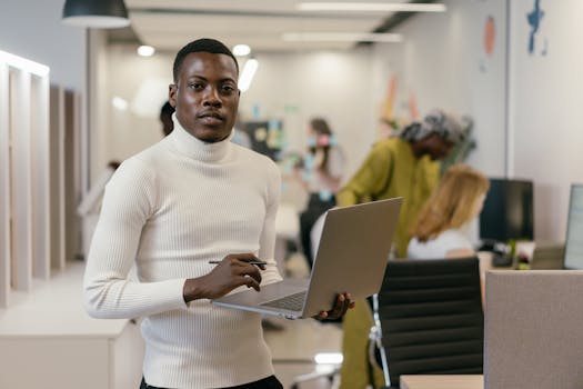 Confident man in white turtleneck working on laptop in a modern office setting.