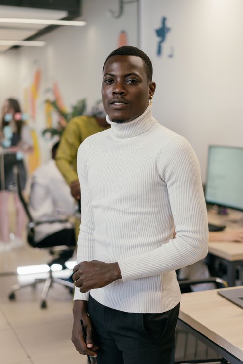 Free A confident African American man in a white turtleneck sweater stands in a modern office. Stock Photo