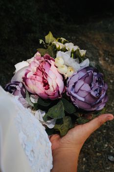 Colorful peony bouquet held in hand, showcasing purple and pink hues with lush green leaves.