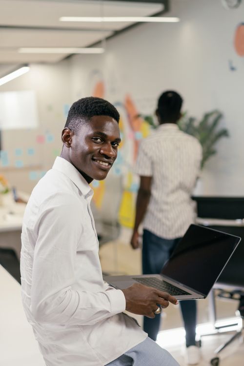 Free A young man in a white shirt smiling while holding a laptop in an office setting. Stock Photo