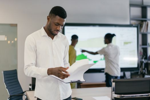 Man in office reading documents with colleagues in the background, strategizing business approaches.