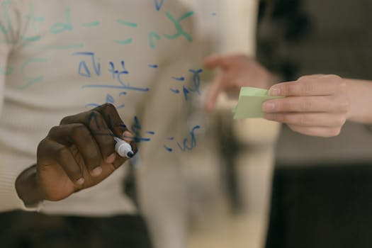 Hands writing formulas on glass board during team brainstorming session.