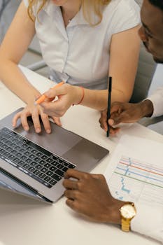 Two colleagues collaborating over a laptop and charts in an office setting.