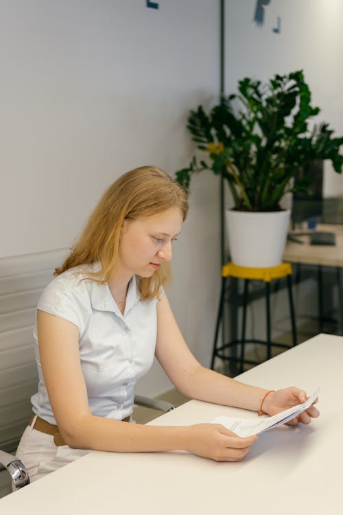 Free Woman in White Button Up Shirt Reading a Piece of White Paper while Sitting on a Chair Stock Photo