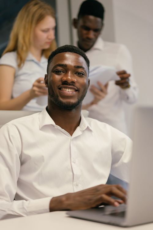 Free Smiling businessman at desk with colleagues in background. Collaborative office setting. Stock Photo