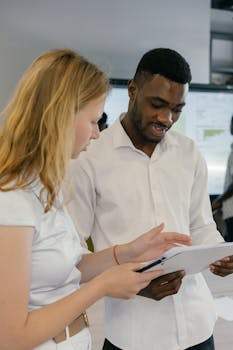 Two young colleagues discussing work in a modern office setting, sharing ideas and reviewing documents.