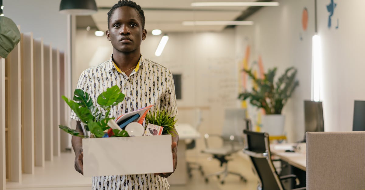 Photo by Mikhail Nilov A serious man carries a box of belongings in an office setting, symbolizing dismissal or job change.