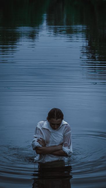 A thoughtful woman stands in calm water at twilight, conveying solitude.