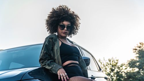 Free Fashionable young woman with sunglasses and curly hair posing by a sleek car outdoors. Stock Photo