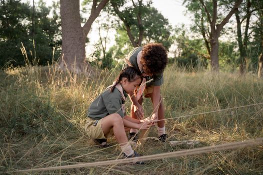 Two young scouts tying knots in a grassy campsite, focused on teamwork and learning.