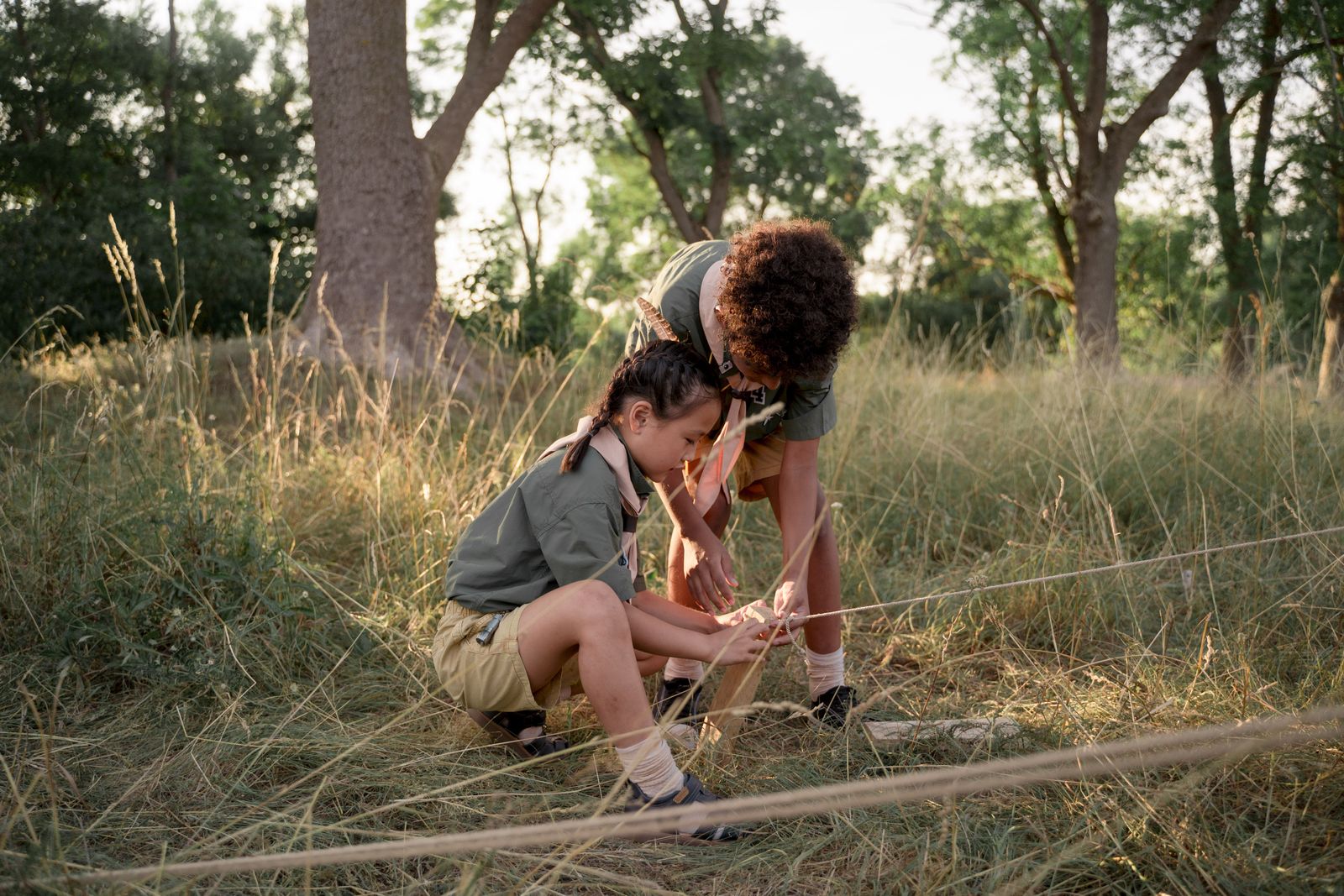 Two young scouts work together tying rope around a wooden stake in a sunny woodland