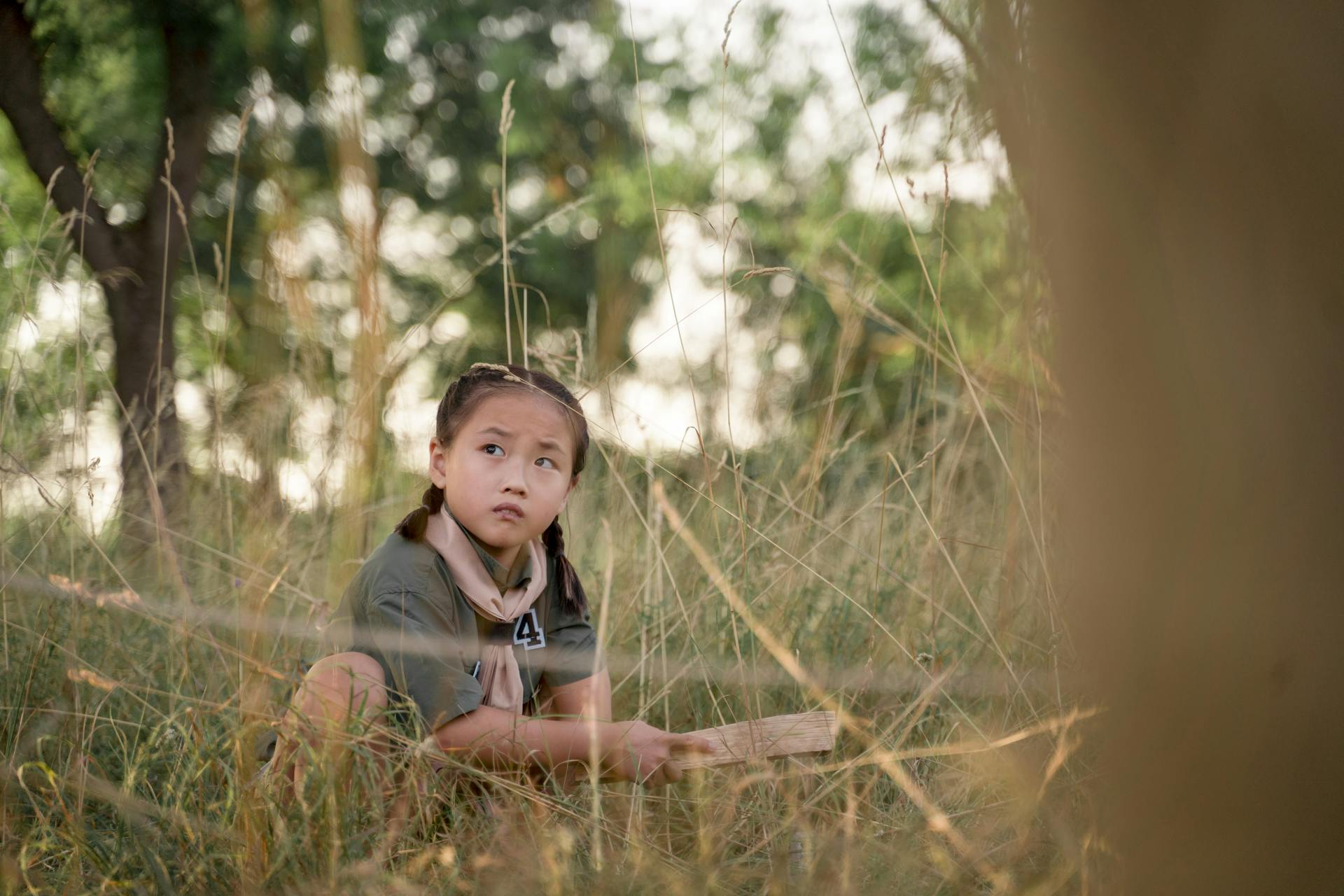 A young girl in a scout uniform crouching in grassy outdoors, engaged in scouting activities.