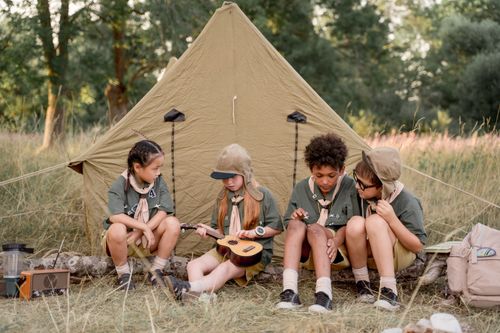 Free A group of children in scout uniforms sitting by a tent playing a ukulele, enjoying the outdoors. Stock Photo