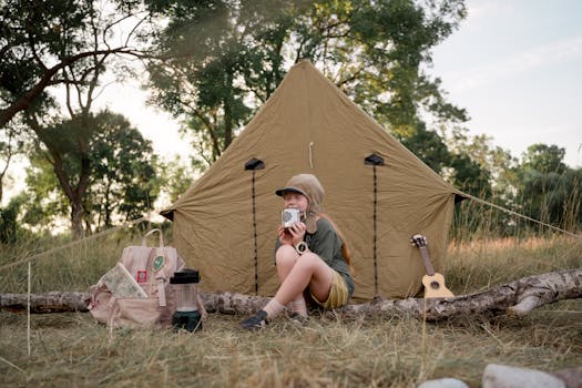 Young camper savoring coffee, sitting beside a tent in the woods with a ukulele.