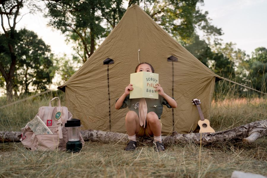Child reading a scout field book at a campsite with a tent, ready for a camping adventure in the forest. Stock Photo