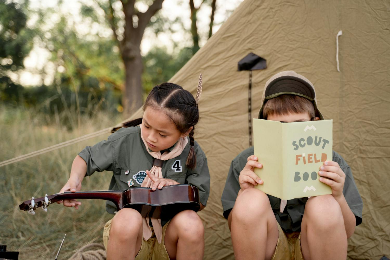 Two young scouts read and play ukulele at an outdoor camp, enjoying nature.