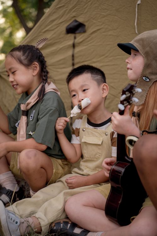 Free Group of children sitting by a tent, having fun with marshmallows and a ukulele at a scout camp. Stock Photo