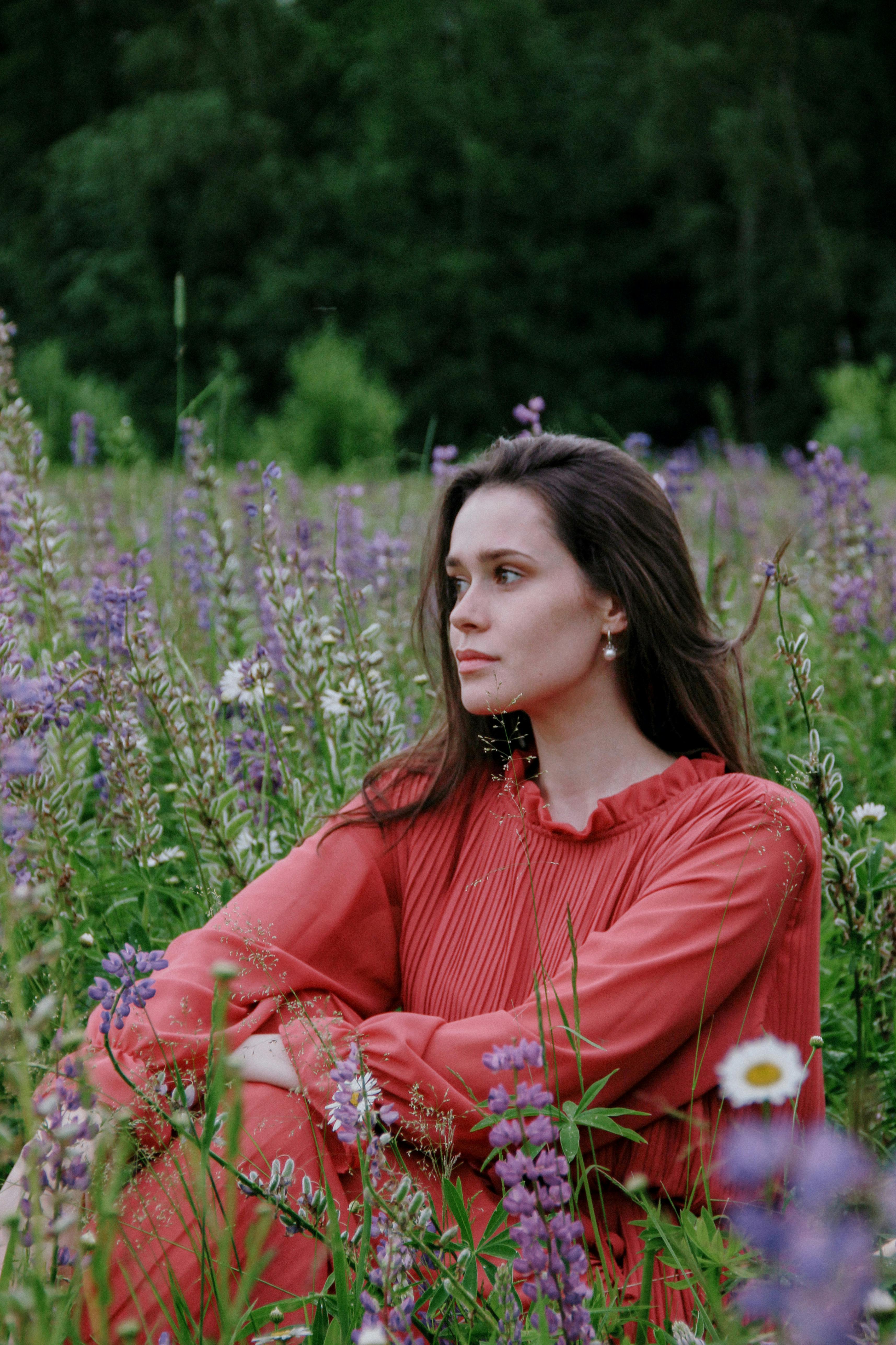 一位穿着红色连衣裙的沉思女子坐在野花丛中，周围是郁郁葱葱的绿色植物