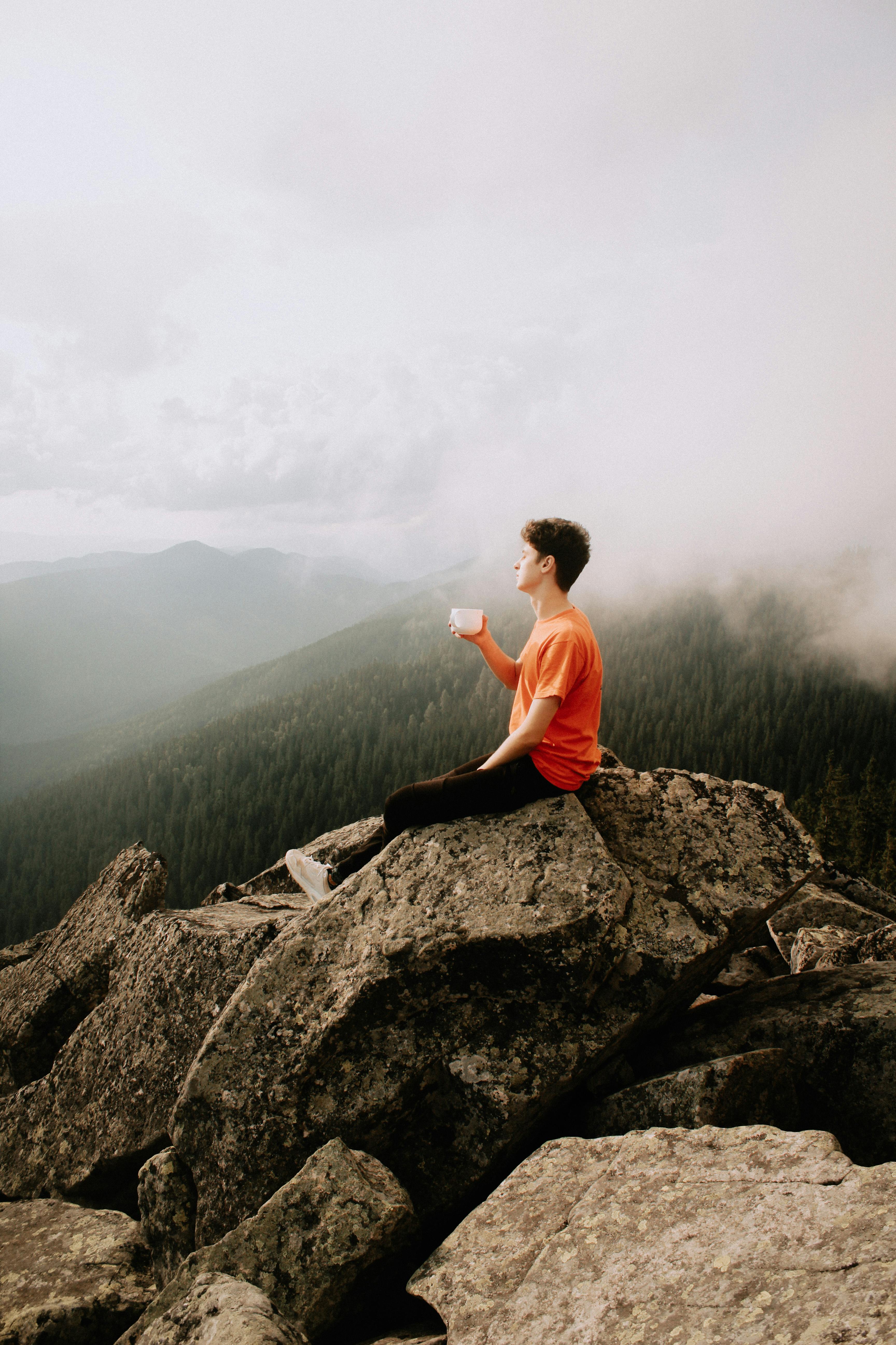 Man relaxing on mountain peak, holding a cup, amidst scenic cloudy landscape.