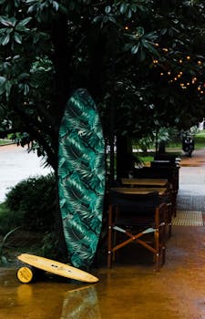 雨水浸湿的街道旁，树灯下摆着户外桌椅，旁边矗立着充满活力的冲浪板