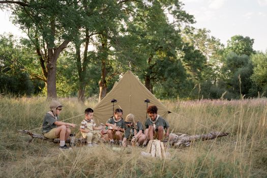 A group of children sitting by a tent in a grassy area, enjoying a camping experience.