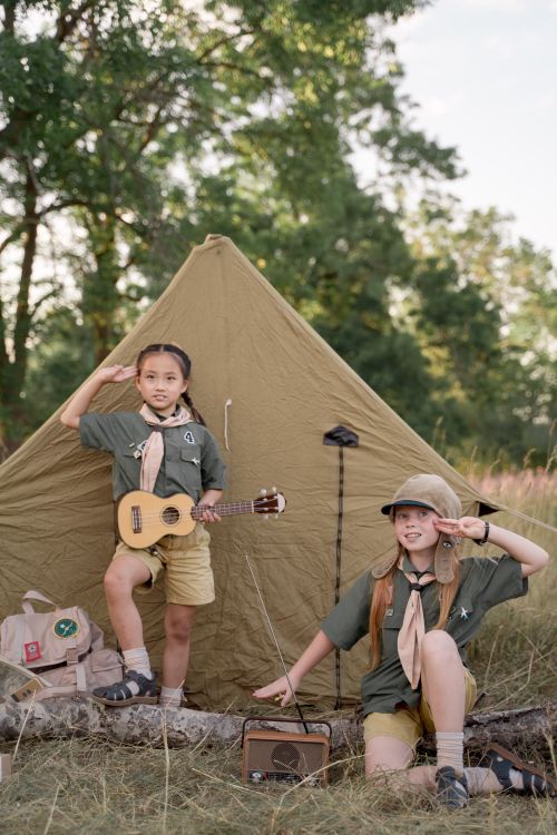 Free Two girls in scout uniforms salute in front of a tent, one holding a ukulele, at a scouting camp. Stock Photo