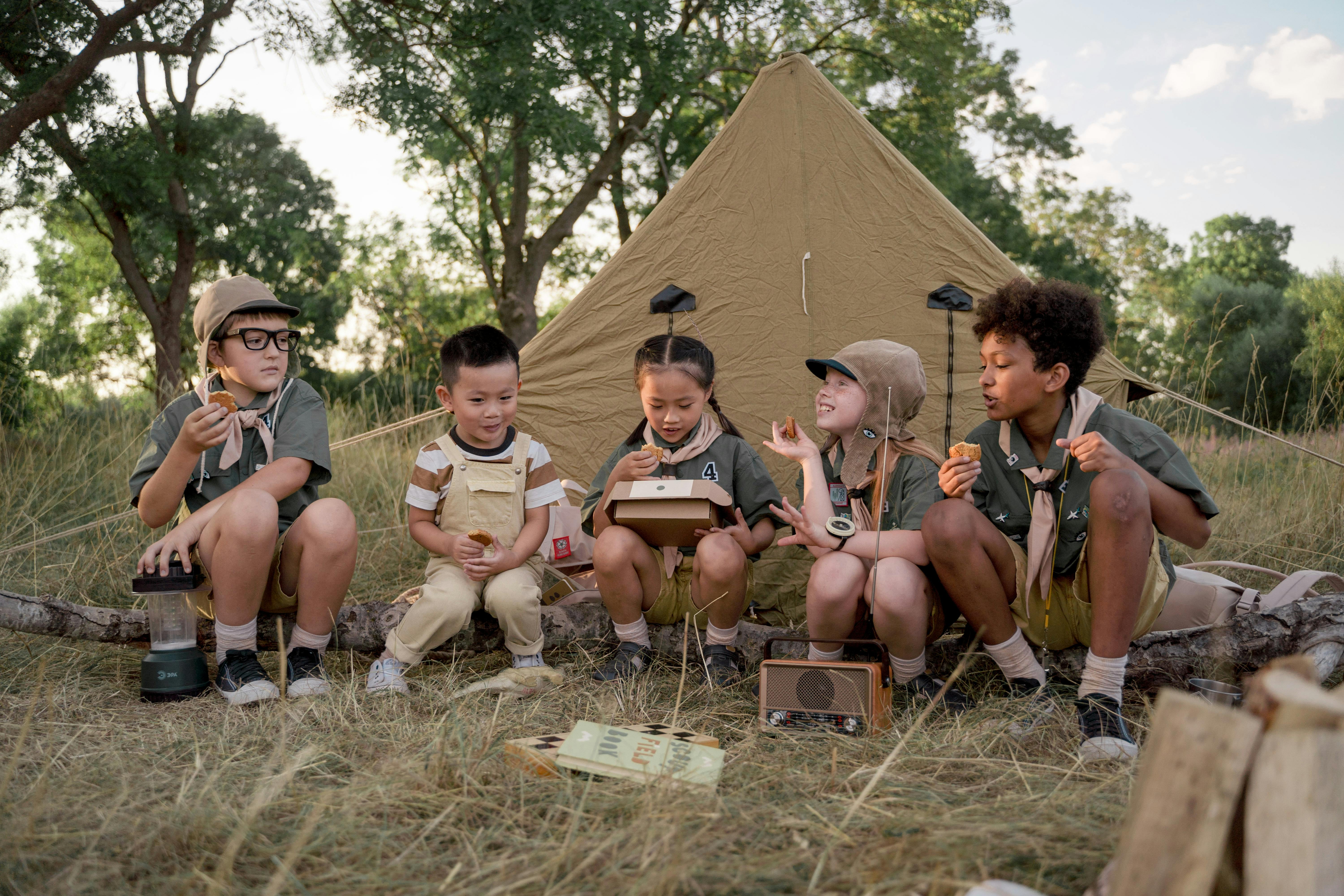 Four children in scout uniforms are seated in front of a tent, smiling and enjoying their outdoor adventure together.