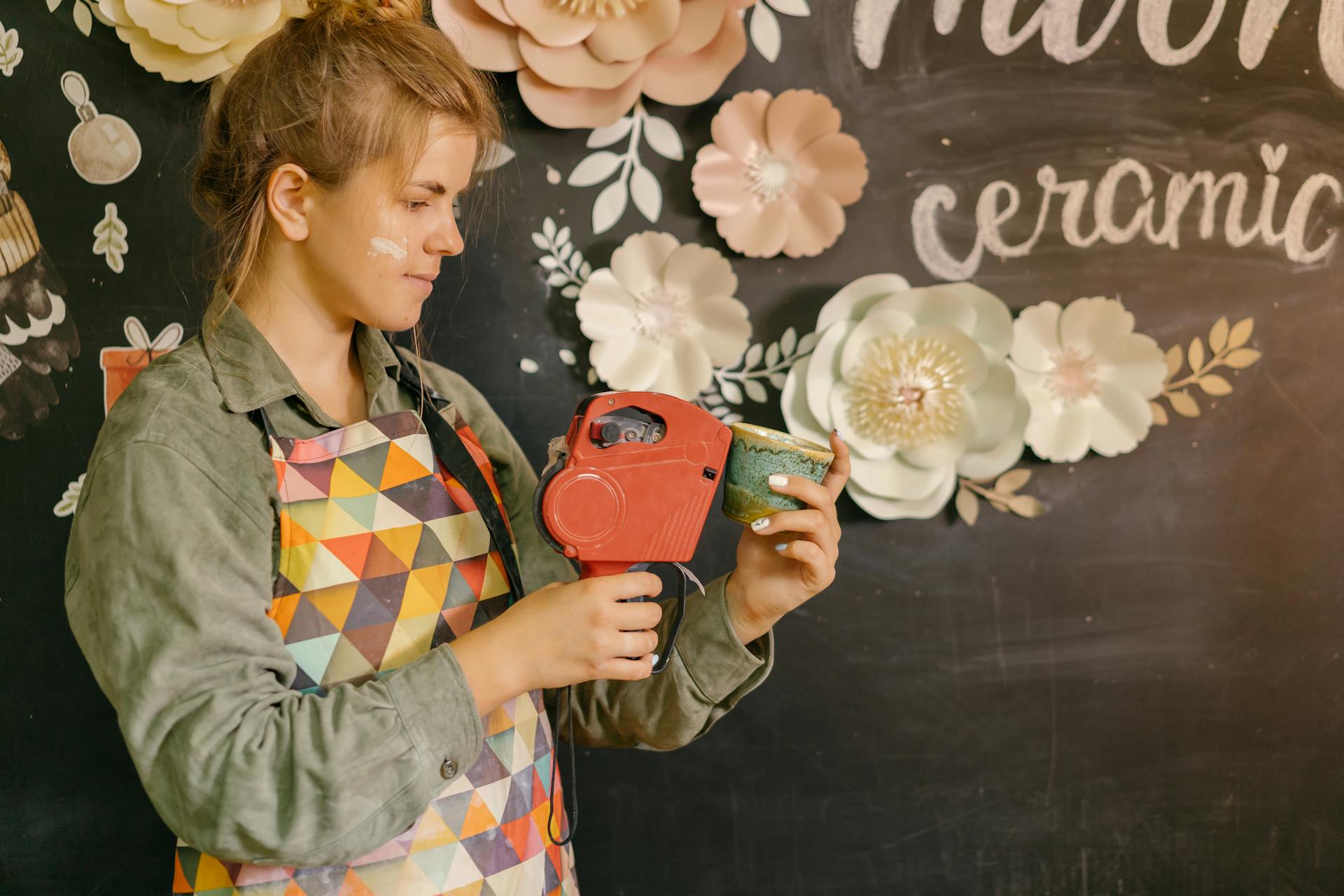 A young woman in an apron labeling a handcrafted ceramic cup in a creative workspace.