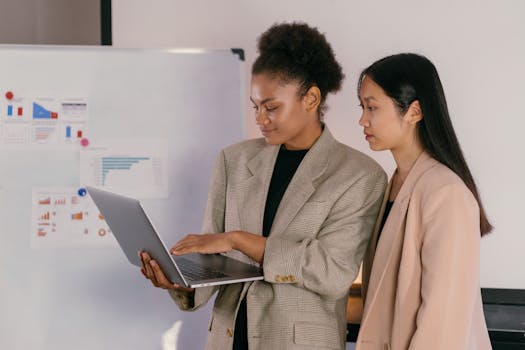 Two professional women discussing data on a laptop in an office setting.