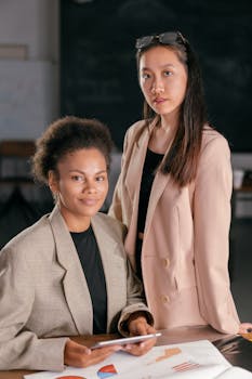 Two businesswomen in blazers confidently posing indoors with office documents.