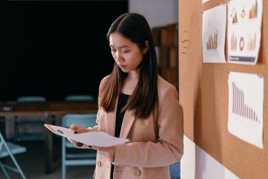 Asian woman in blazer reviewing paper reports with charts in office setting.