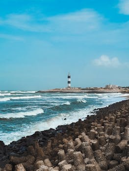 Stunning shot of a lighthouse with waves crashing on a rocky seashore under a bright blue sky.