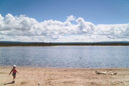 Free A child in a swimsuit walks along a sandy beach by a calm lake under a cloudy sky. Stock Photo