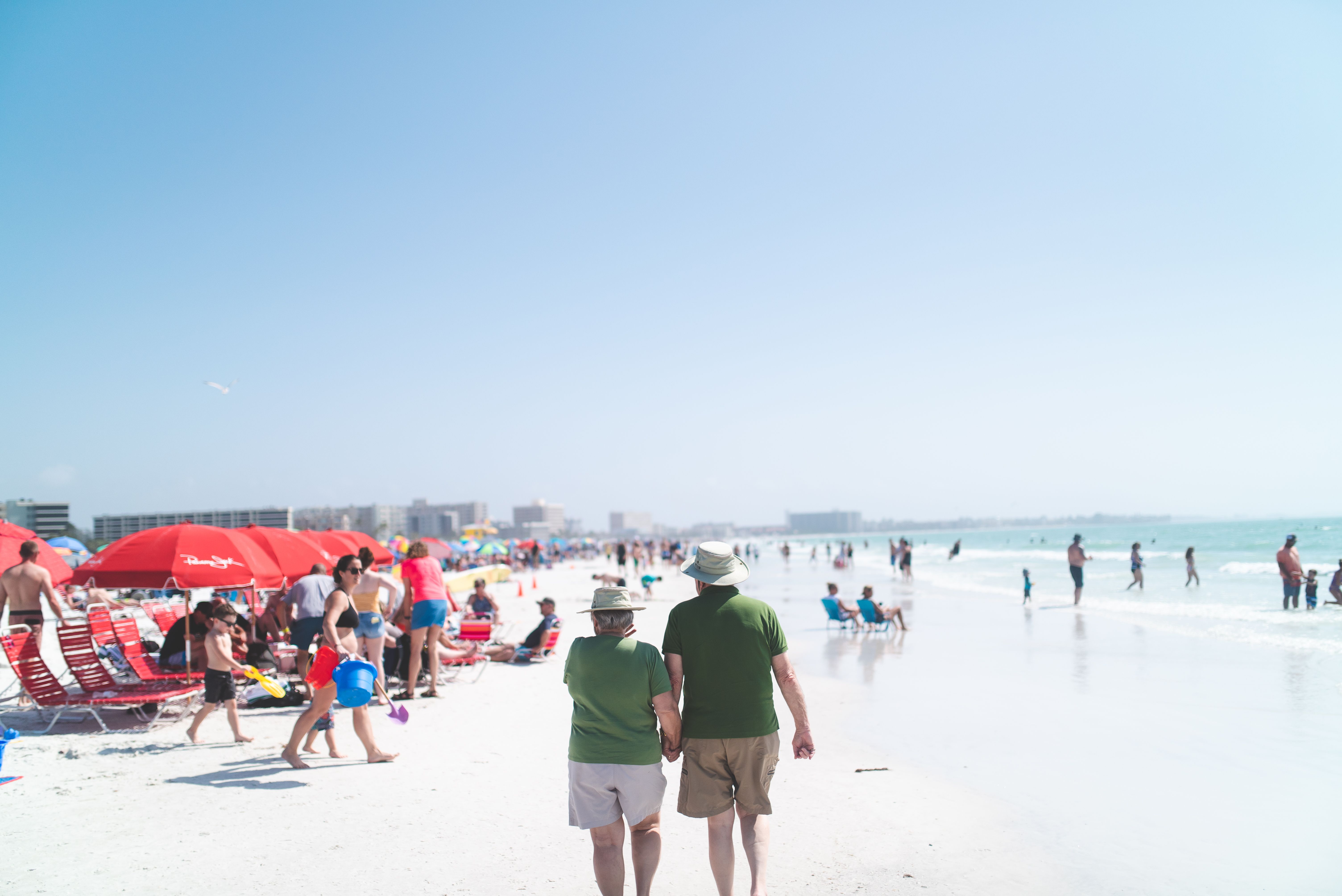 Free A senior couple enjoying a sunny day on a crowded beach, walking hand in hand along the shore. Stock Photo