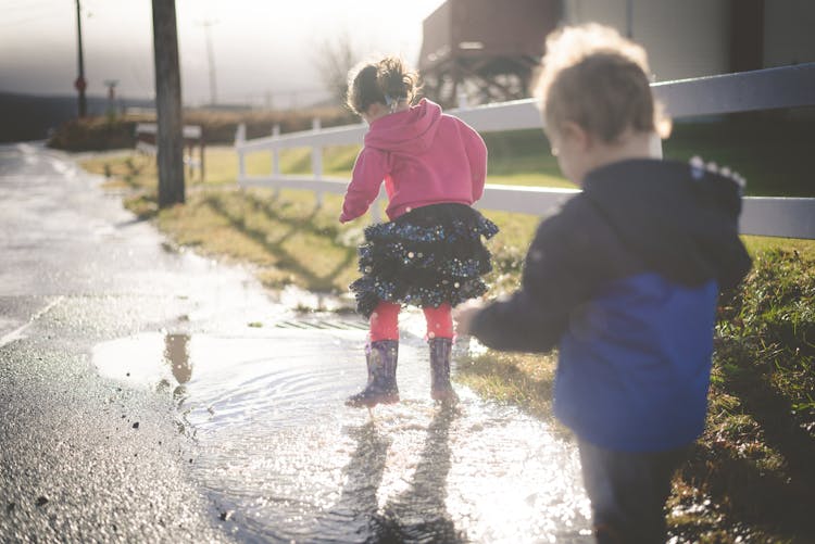 Little Boy And Girl Jumping In A Paddle In Wellies 