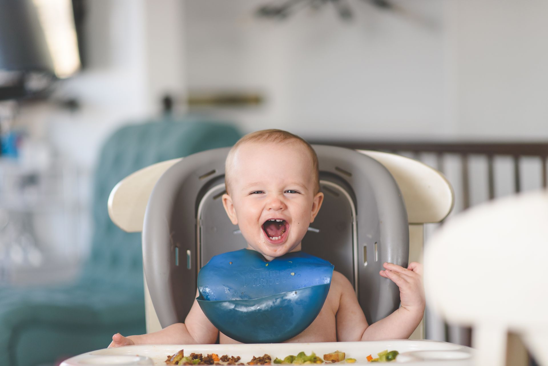 Happy baby laughing in a high chair during mealtime, wearing a blue bib.