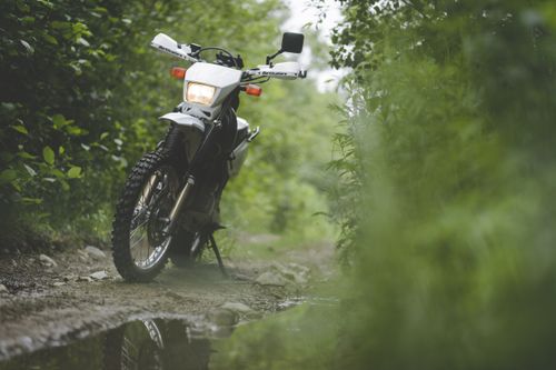 Free White motorbike parked on a muddy path in a rural area surrounded by greenery. Stock Photo